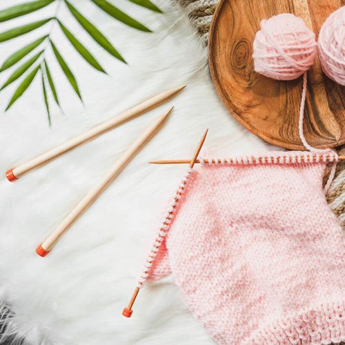Knitting needles and pink yarn on a wooden surface with green leaves.