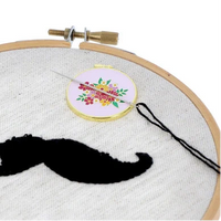Embroidery hoop with a black mustache embroidery, and a small floral needle minder on a white background