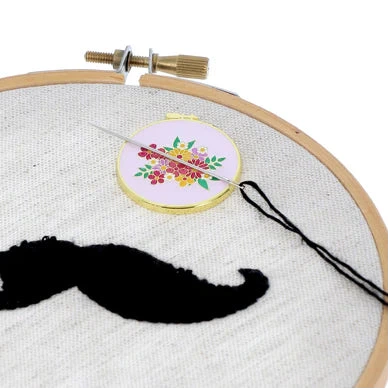 Embroidery hoop with a black mustache embroidery, and a small floral needle minder on a white background