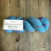 A skein of Cascade Yarn220 Superwash Sport Wave yarn in a variegated blue, teal, and pink colorway, resting on a rustic wooden surface with its paper label visible.