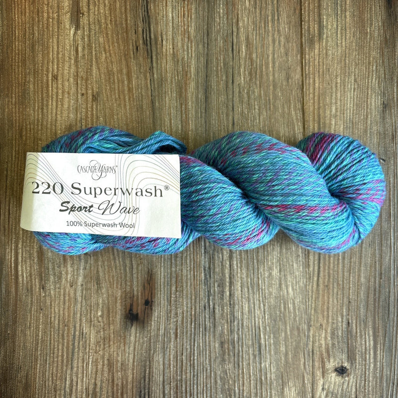 A skein of Cascade Yarn220 Superwash Sport Wave yarn in a variegated blue, teal, and pink colorway, resting on a rustic wooden surface with its paper label visible.
