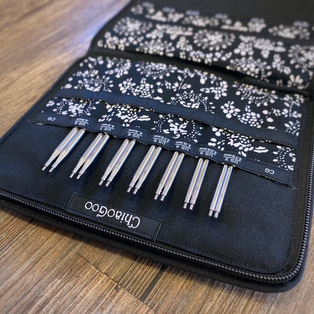 Set of metal knitting needles in a black case with floral pattern on a wooden surface