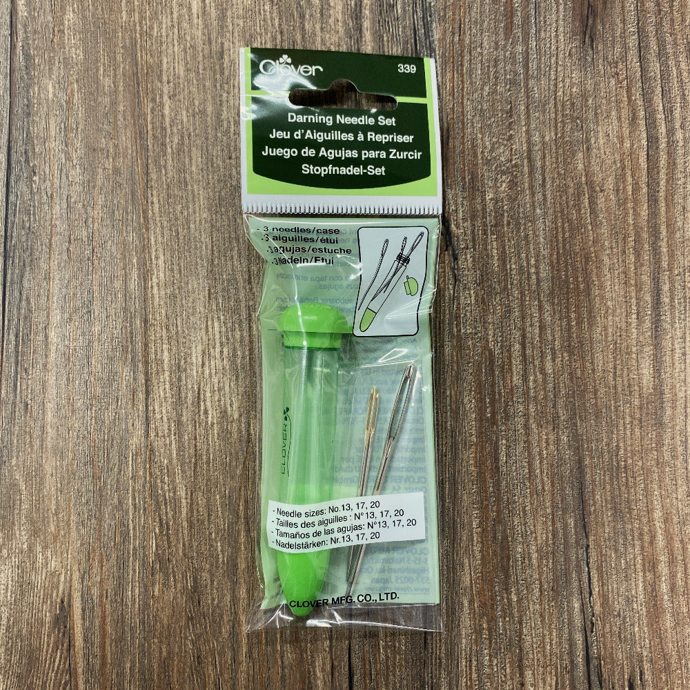 A packaged darning needle set in clear plastic with a green and white header card, photographed on a wood surface. The set includes several metal darning needles and a green stopper in a clear tube.