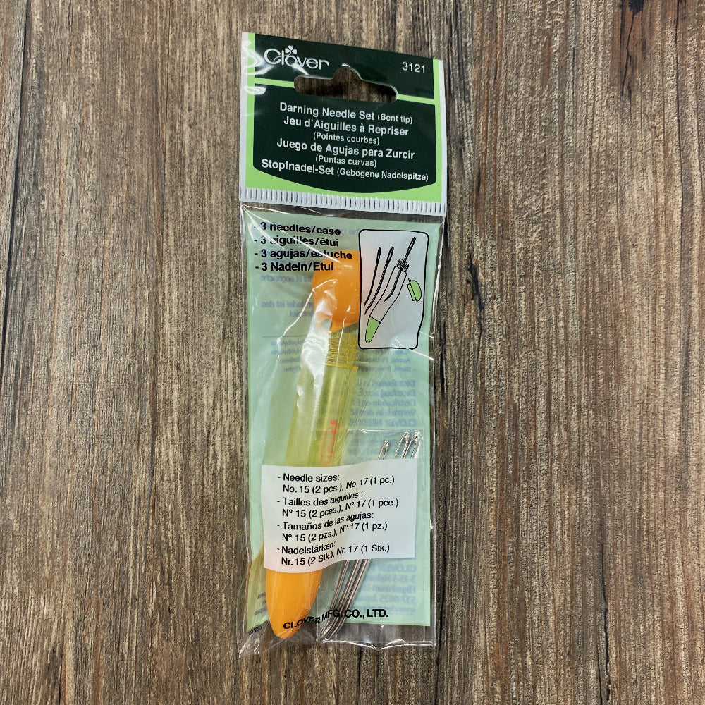 A packaged darning needle set in clear plastic packaging with a green and black header card, placed on a brown wood surface. The set includes several metal needles and an orange container