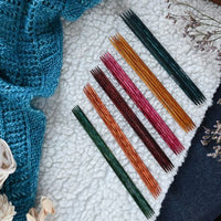 Colorful double pointed knitting needles on a textured surface with a blue knitted fabric and dried flowers.