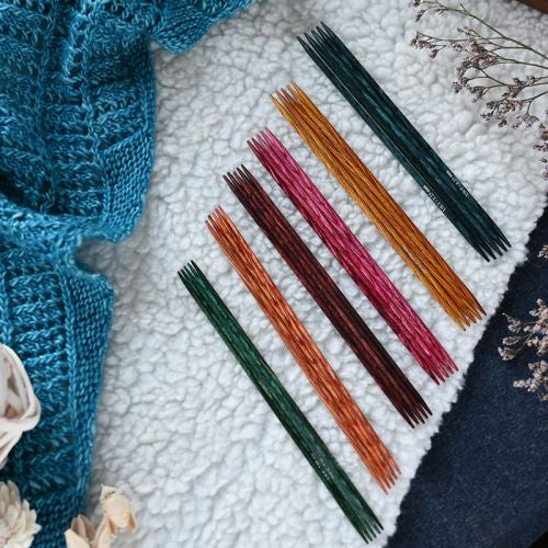 Colorful double pointed knitting needles on a textured surface with a blue knitted fabric and dried flowers.