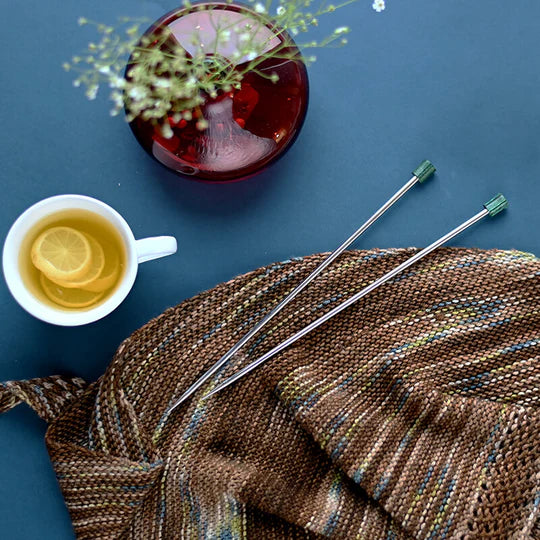 A pair of silver and green knitting needles on a brown knit project surrounded by a teacup and decorative accents on a blue background.