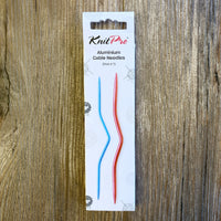 White KnitPro box with 2 sizes of cable needles on it, on a wood background