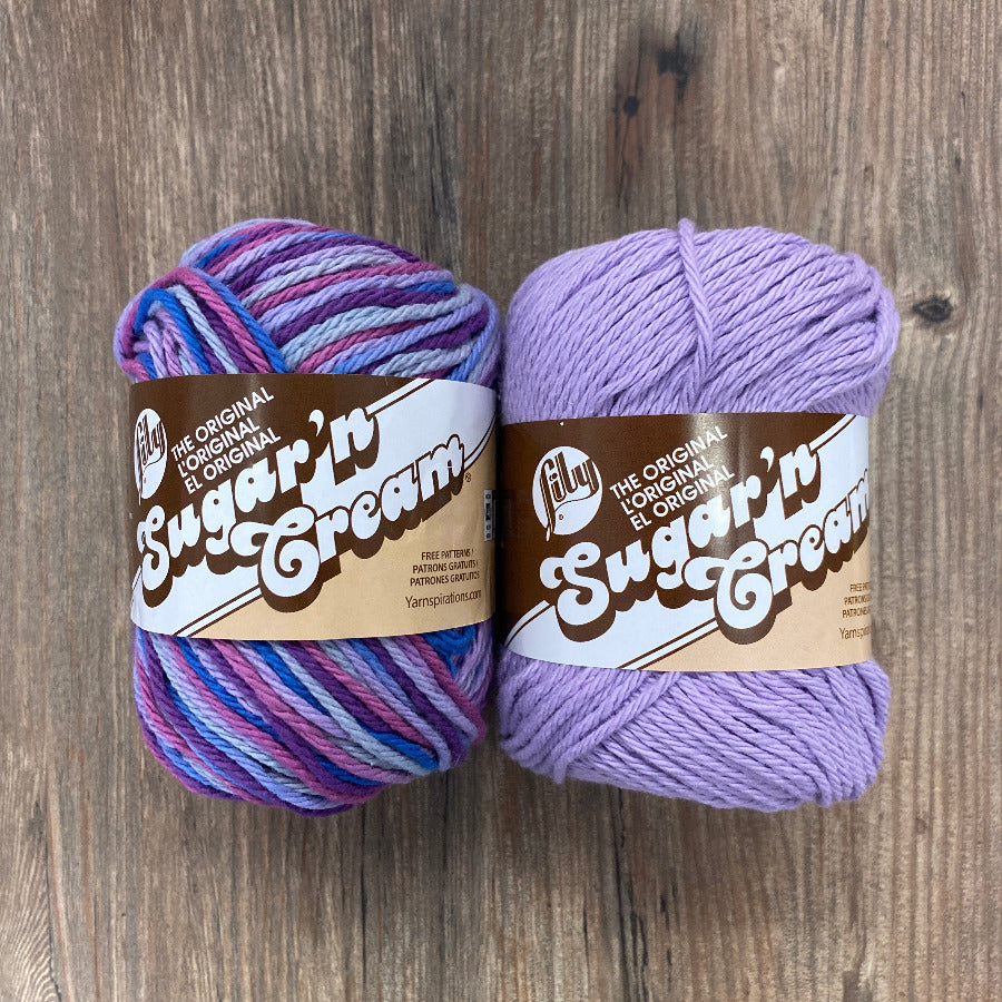 Two skeins of yarn, one multicolored and one purple, on a wooden surface with 'Sugar'n Cream' labels.