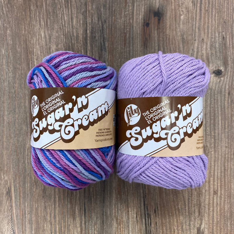 Two skeins of yarn, one multicolored and one purple, on a wooden surface with 'Sugar'n Cream' labels.