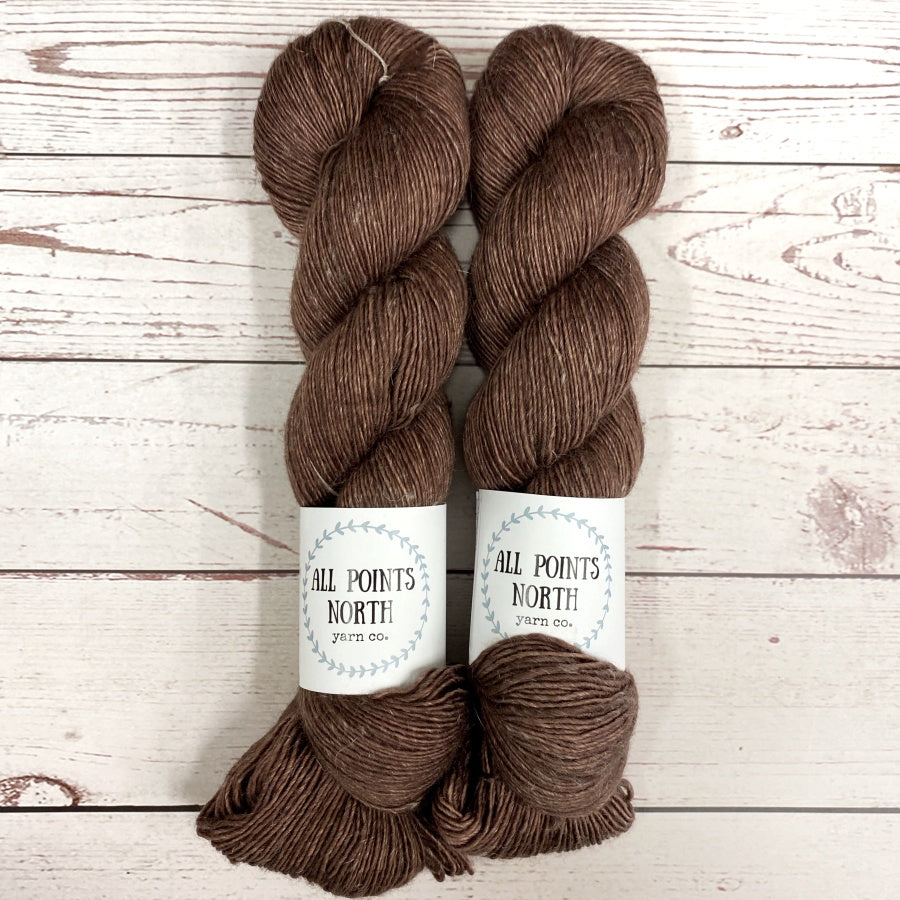 Two skeins of brown yarn with All Points North labels on a white wood background
