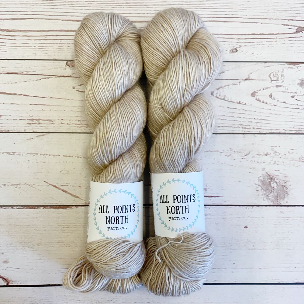 Two skeins of beige yarn with All Points North labels on a white wood background