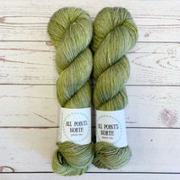 Two skeins of olive yarn with All Points North labels on a white wood background