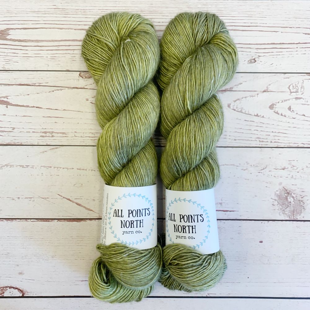 Two skeins of olive yarn with All Points North labels on a white wood background