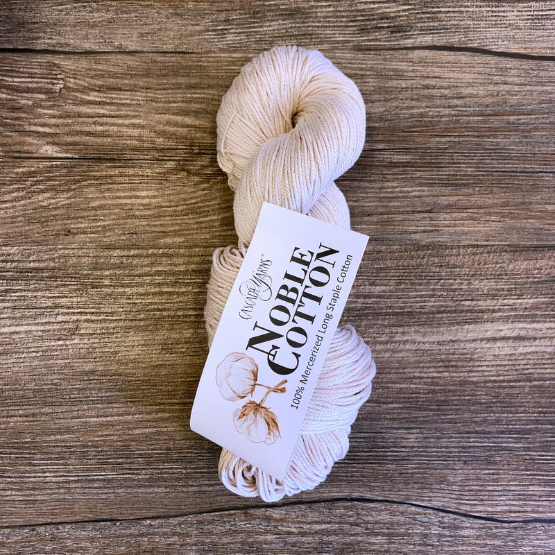 Ball of cream-colored yarn with a 'Noble Cotton' label on a wooden surface