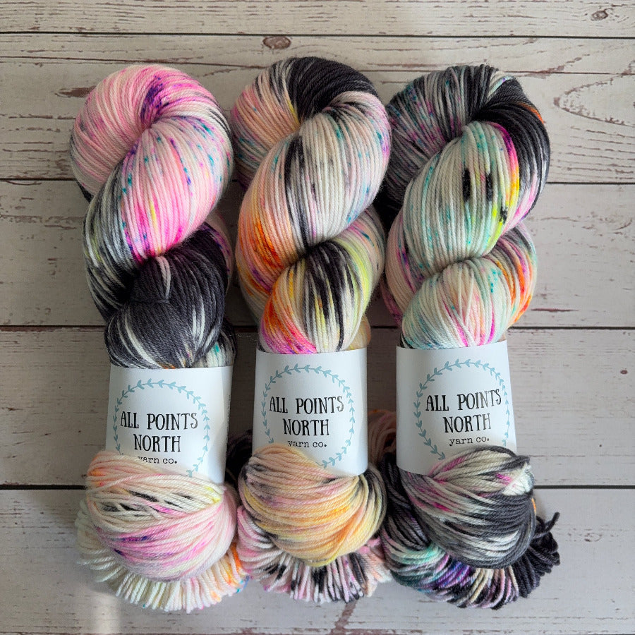 Three skeins of variegated black, white, and fluorescent speckled yarn from All Points North Yarn Co. arranged side-by-side on a light whitewashed wooden background.
