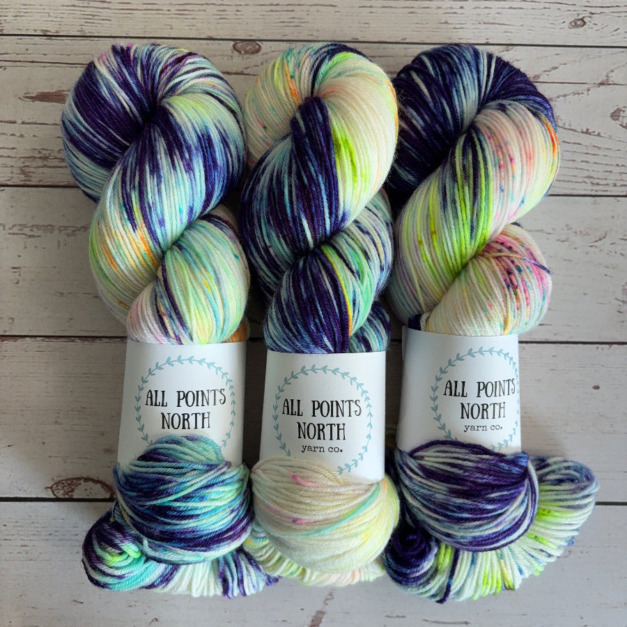 Three skeins of variegated blue and white with fluorescent speckles yarn from All Points North Yarn Co. arranged side-by-side on a light wood background.