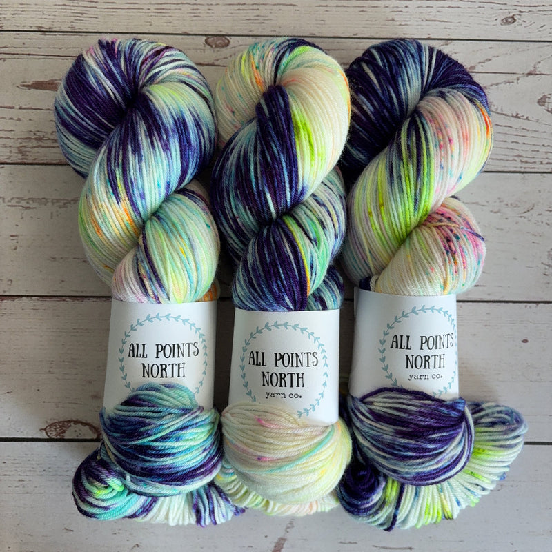 Three skeins of variegated blue and white with fluorescent speckles yarn from All Points North Yarn Co. arranged side-by-side on a light wood background.