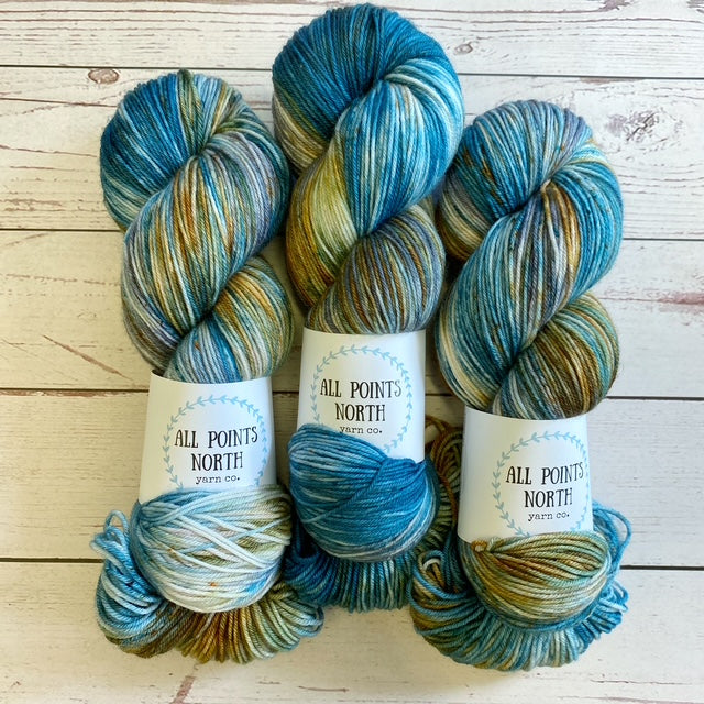 Three blue, rust, grey, and white yarn skeins with an All Points North label on a white wood background