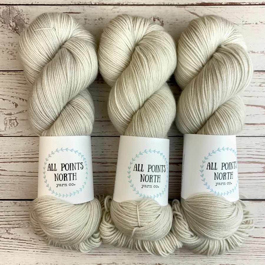 Three skeins of yarn in a soft off-white/creamy grey shade, each with a white paper label reading 'ALL POINTS NORTH yarn co.' placed on a whitewashed wood surface.