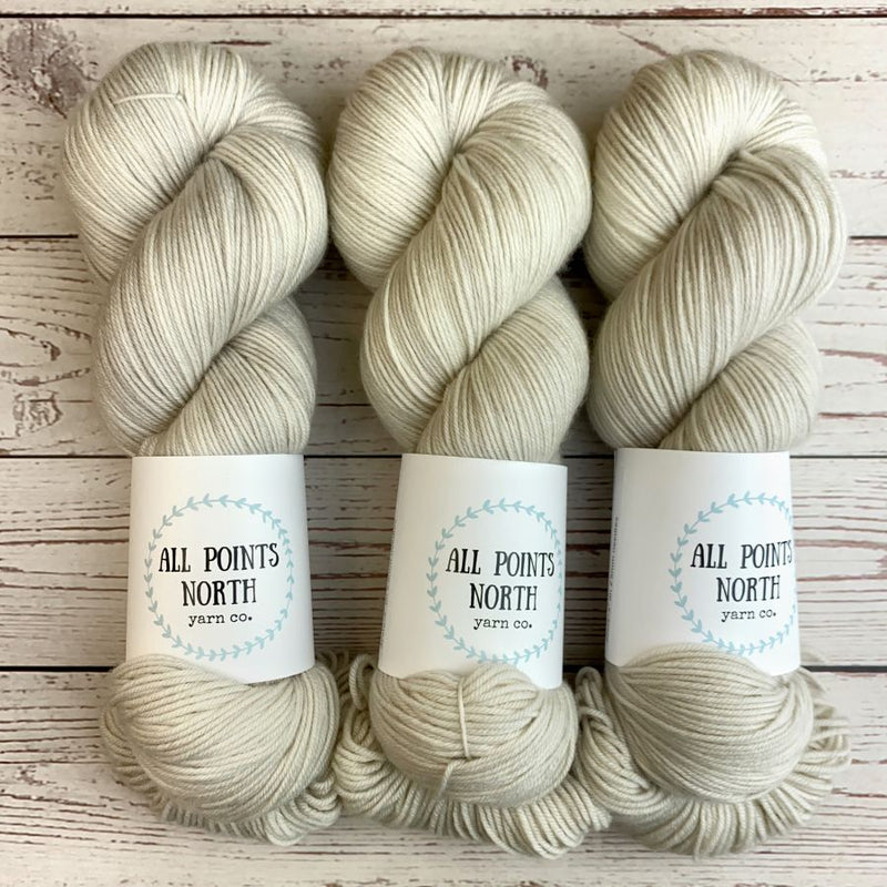 Three skeins of yarn in a soft off-white/creamy grey shade, each with a white paper label reading 'ALL POINTS NORTH yarn co.' placed on a whitewashed wood surface.