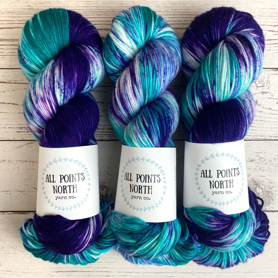 Three hanks of variegated purple and aqua yarn with white paper labels reading 'ALL POINTS NORTH yarn co.' arranged side by side on a light wood background.