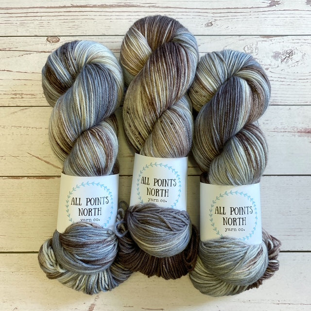 Three grey, brown, and white yarn skeins with an All Points North label on a white wood background