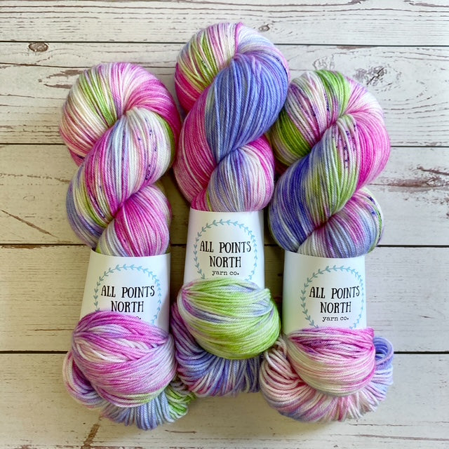 Three multicoloured pink, white, purple, and green yarn skeins with an All Points North label on a white wood background