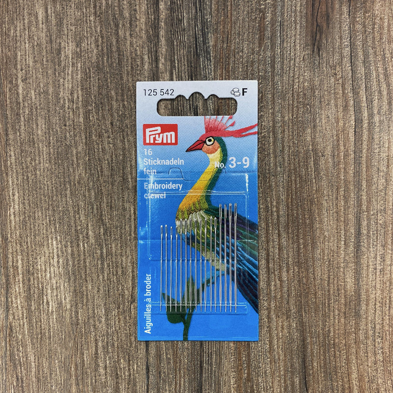 Pack of embroidery needles with a colorful bird design on the packaging against a wooden background
