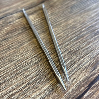 Two stainless steel Quads interchangeable needle tips, placed diagonally on a wood-grain surface.