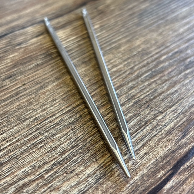 Two stainless steel Quads interchangeable needle tips, placed diagonally on a wood-grain surface.