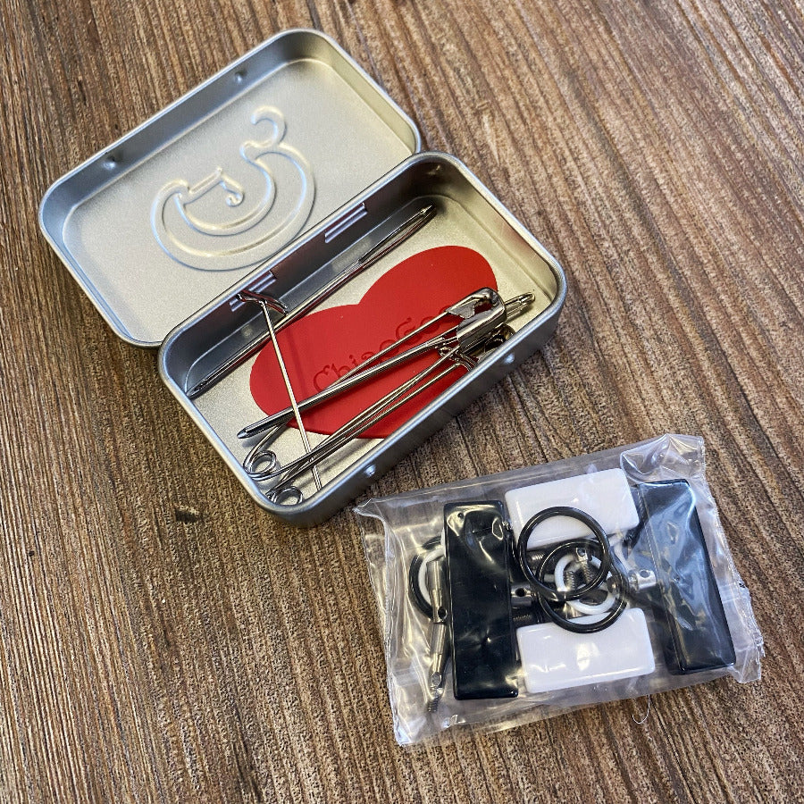 Open metal tin with safety pins and a clear plastic bag filled with interchangeable needle accessories on a wooden surface