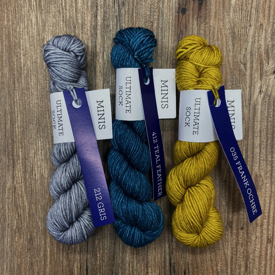 Three mini skeins of yarn in gray, blue, and yellow with labels on a wooden surface